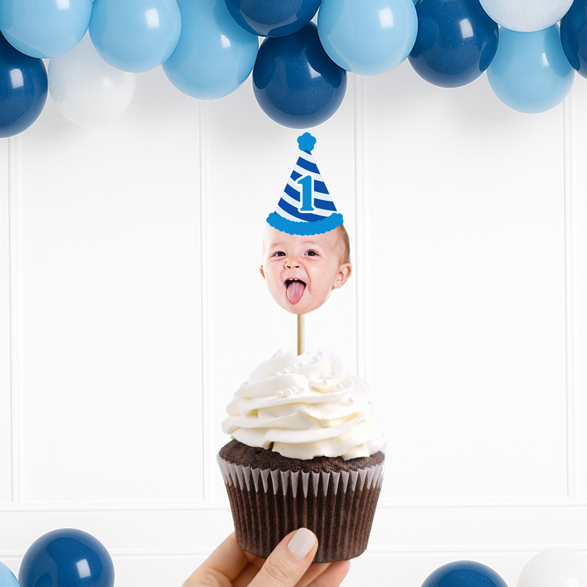 A custom birthday party cupcake topper featuring a baby's face sticking out of a frosted chocolate cupcake, topped with a blue and white striped first birthday hat. The hand holding the cupcake is shown against a white paneled wall decorated with blue and white balloons.