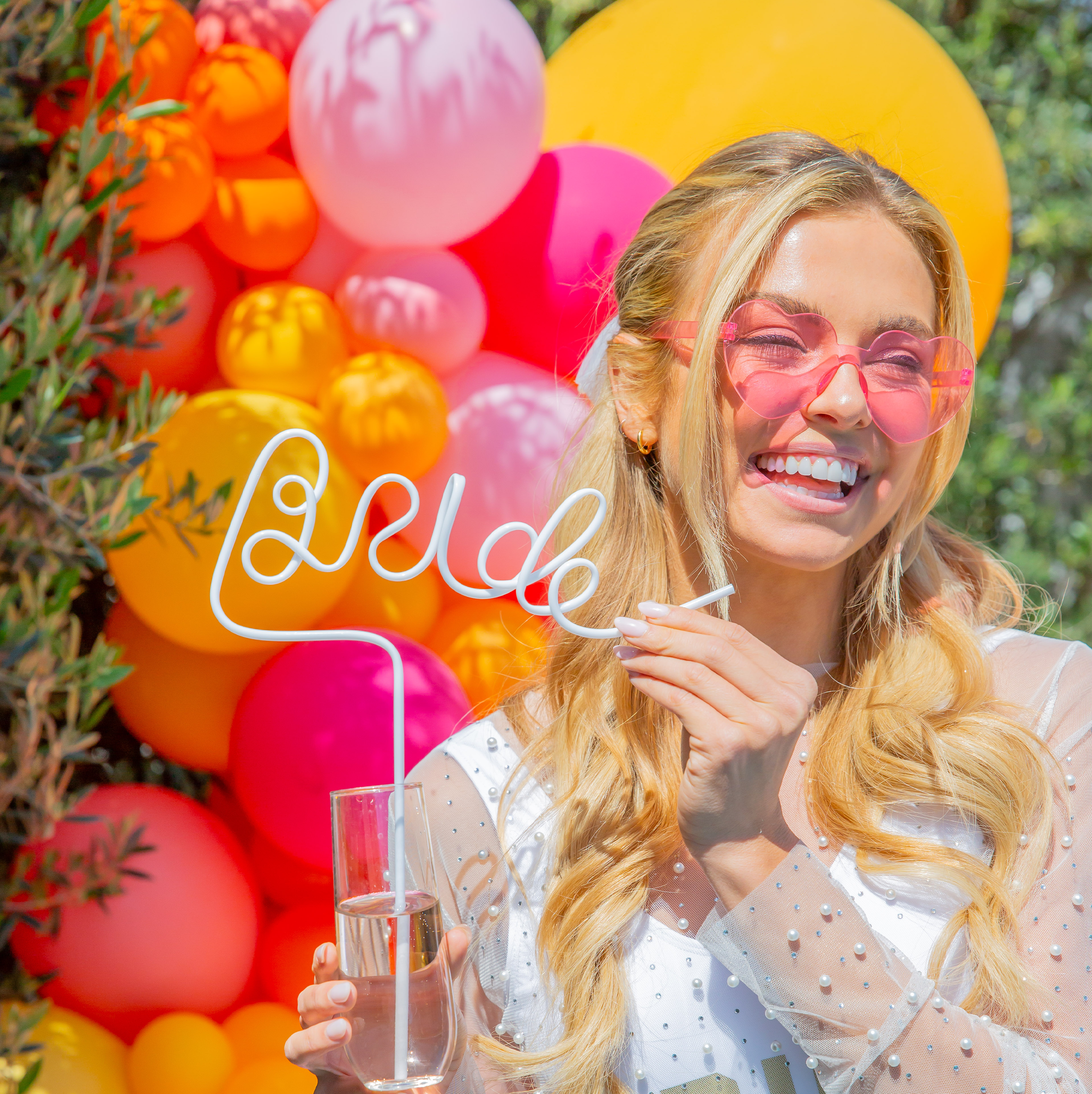 Bright balloon background with the bride holding a white bride silly straw and wearing Sunkissed Party Co. heart sunglasses at her bachelorette.