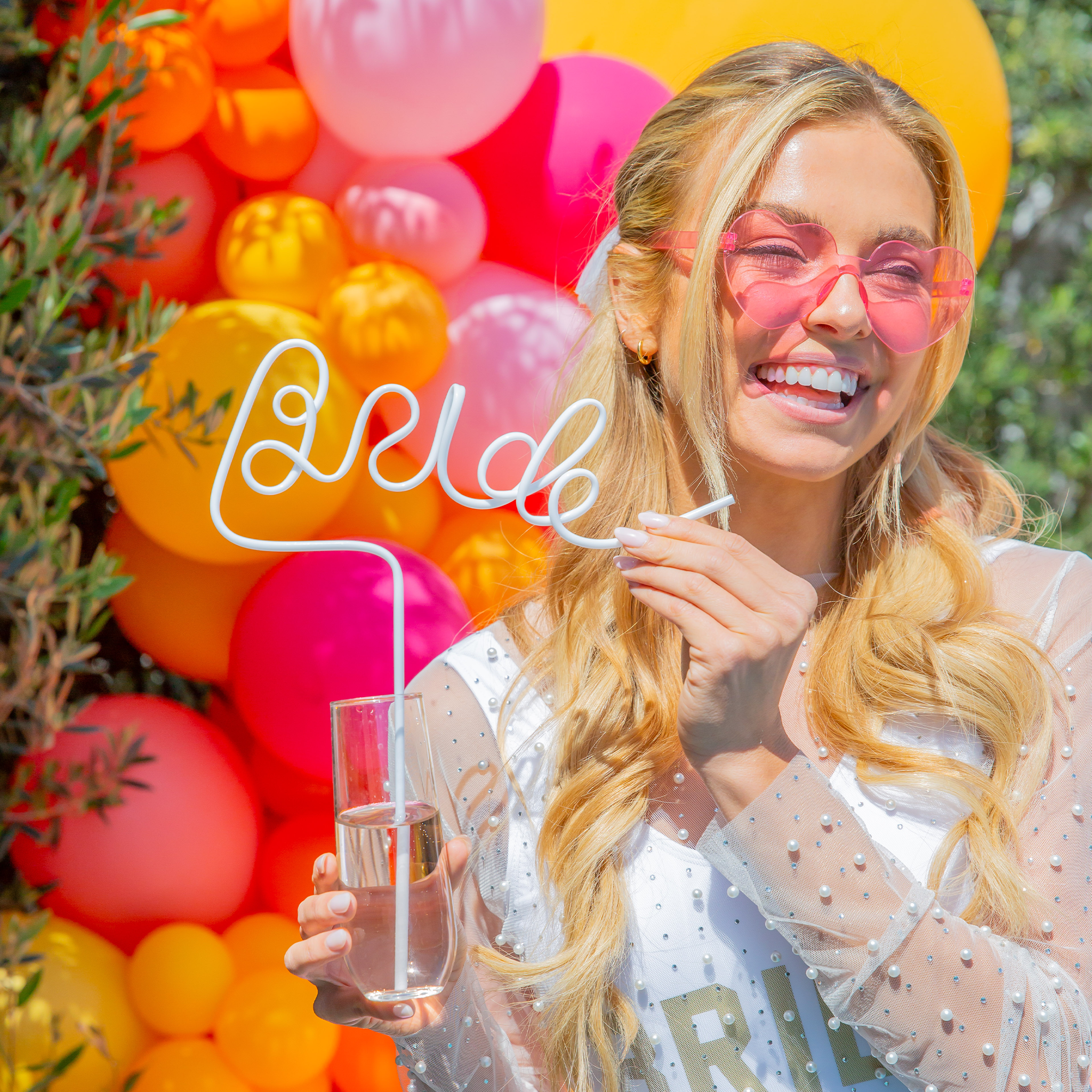 Bride at bachelorette party holding a white Bride straw, wearing pearl veil and heart sunglasses, with balloon arch backdrop.